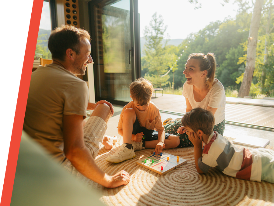 Family game night: parents and two children playing a board game on rug by open sliding door in sunlit home.