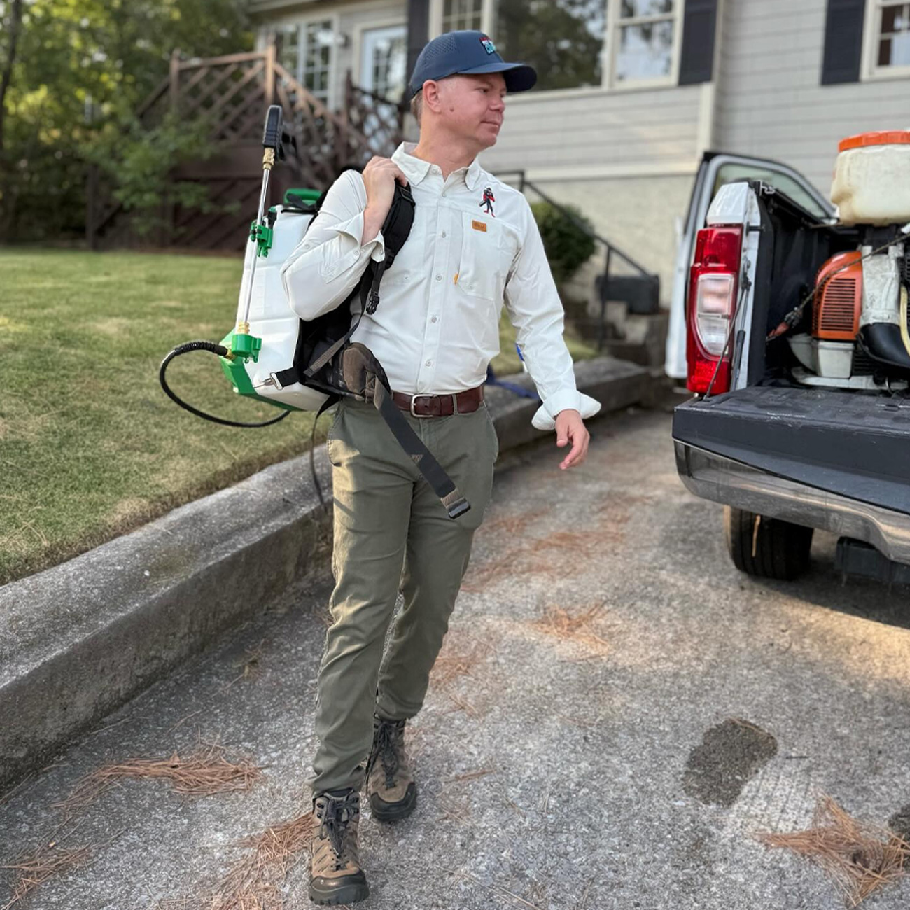 Pest control technician carrying backpack sprayer walking to pickup truck beside house